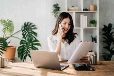 İş kadını Asyalı kadın telefonda konuşuyor ve ev ofisinde laptop kullanıyor.