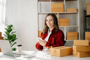 Startup small business SME, Entrepreneur owner woman using laptop taking receive and checking online purchase shopping order to prepare pack product boxes.  at home office clipart