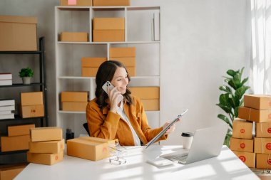 Başlangıçta küçük işletme KOBİ 'si, dizüstü bilgisayarı kullanan girişimci kadın paket ürün kutuları hazırlamak için online alışveriş siparişlerini alıp kontrol ediyor. ev ofisinde.