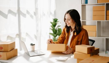 Başlangıçta küçük işletme KOBİ 'si, dizüstü bilgisayarı kullanan girişimci kadın paket ürün kutuları hazırlamak için online alışveriş siparişlerini alıp kontrol ediyor. ev ofisinde.
