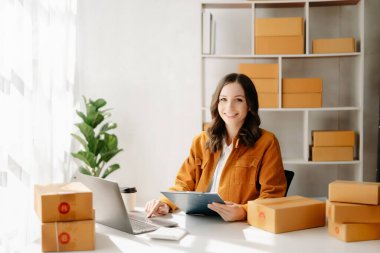 Başlangıçta küçük işletme KOBİ 'si, dizüstü bilgisayarı kullanan girişimci kadın paket ürün kutuları hazırlamak için online alışveriş siparişlerini alıp kontrol ediyor. ev ofisinde.