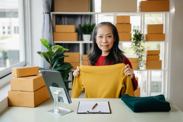 Fashion blogger concept, Senior Asian woman selling clothes on video streaming.Startup small business SME, using tablet taking receive and checking at home office
