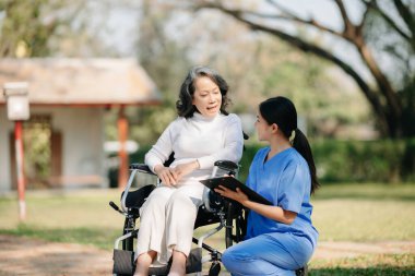 Asyalı yaşlı, yaşlı, yaşlı, tekerlekli sandalyede, Asyalı, dikkatli bir bakıcı ve bahçede yürüyen bir hastayı cesaretlendiren bir kadın. Bakıcının ve kıdemli sağlık sigortasının dikkatine.