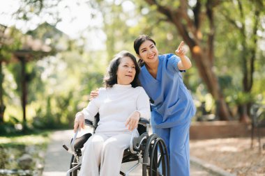 Asyalı yaşlı, yaşlı, yaşlı, tekerlekli sandalyede, Asyalı, dikkatli bir bakıcı ve bahçede yürüyen bir hastayı cesaretlendiren bir kadın. Bakıcının ve kıdemli sağlık sigortasının dikkatine..