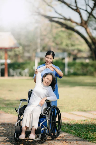 Asian careful caregiver or nurse and the happy patient in a wheelchair are walking in the garden to help and encourage and rest your mind with green nature. Help support yourself to learn to walk.