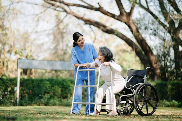 Asian careful caregiver or nurse and the happy patient with wheelchair are walking in the garden to help and encourage and rest your mind with green nature. Help support yourself to learn to walk.