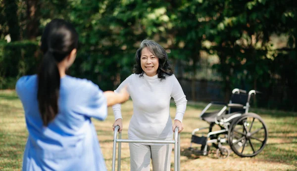 stock image Asian careful caregiver or nurse and the happy patient with wheelchair are walking in the garden to help and encourage and rest your mind with green nature. Help support yourself to learn to walk. walker in sun ligh