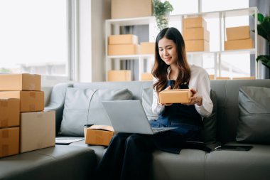 Genç işletme sahibi kadın, paket kutuları hazırla ve dizüstü bilgisayardaki müşteriye teslim edilmesi için online siparişleri kontrol et. 