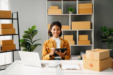 Başlangıçta küçük işletme KOBİ 'si, tablet ve laptop kullanan girişimci Afrikalı kadın paket ürün kutularını hazırlamak için online alışveriş siparişi alıyor ve kontrol ediyor. Merkez ofiste.