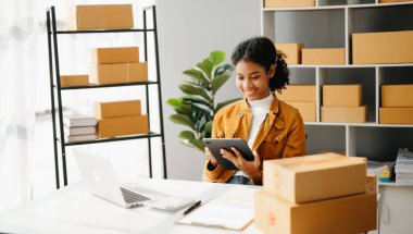 Başlangıçta küçük işletme KOBİ 'si, tablet ve laptop kullanan girişimci Afrikalı kadın paket ürün kutularını hazırlamak için online alışveriş siparişi alıyor ve kontrol ediyor. Merkez ofiste.