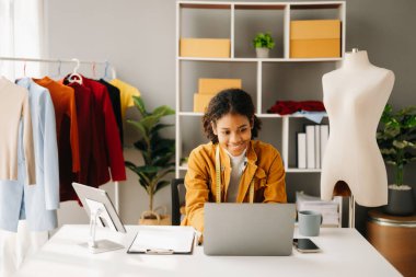 Sakin, kıvırcık esmer, koyu tenli kadın modern moda tasarımcısı, dizüstü bilgisayar ve tablet bilgisayarın ofisinde.