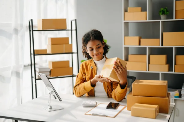 stock image Startup small business SME, Entrepreneur owner African woman using  tablet taking receive and checking online purchase shopping order to prepare pack product boxes. in home office