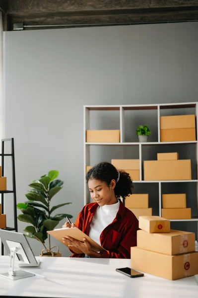 stock image Startup small business SME, Entrepreneur owner African woman using  tablet taking receive and checking online purchase shopping order to prepare pack product boxes. in home office