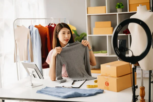 Fashion blogger concept, Young Asian woman selling clothes on video streaming.Startup small business SME, using smartphone and tablet taking receive and checking in office
