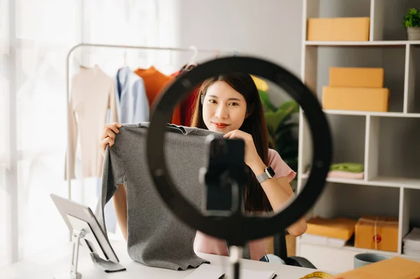 Fashion blogger concept, Young Asian woman selling clothes on video streaming.Startup small business SME, using smartphone or tablet taking receive and checking in office