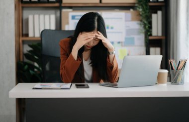 Asyalı kadın migren ağrısı çekiyor. Modern ofiste dizüstü bilgisayarı olan aşırı çalışan iş kadını finansör..