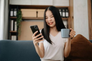 Asyalı kadın evdeki kanepede akıllı telefon kullanıyor.