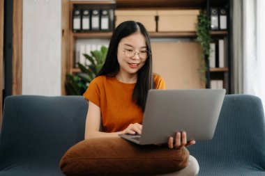  Kanepede oturan Asyalı kız oturma odasında dizüstü bilgisayar kullanıyor.