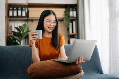  Kanepede oturan Asyalı kız oturma odasında dizüstü bilgisayar kullanıyor.