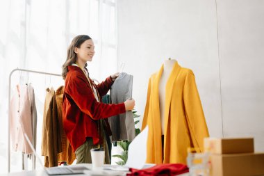 Asyalı terzi kadın terzi atölyesinde giysiler üzerinde çalışıyor. Güzel genç bayan moda tasarımcısı ofiste.