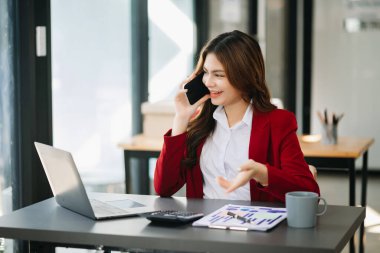 İş kadını Asyalı kadın telefonda konuşuyor ve ofiste otururken bilgisayarını gülümsüyor.