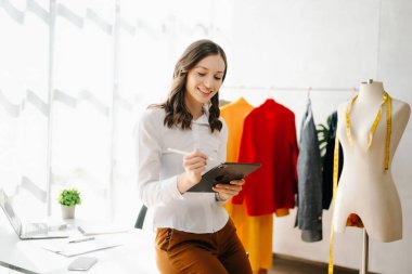 Akıllı telefonlu moda tasarımcısı kadın ve modern stüdyoda dijital tablet bilgisayarlı dizüstü bilgisayar kullanıyor. 