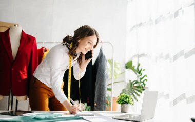 Akıllı telefonlu moda tasarımcısı kadın ve modern stüdyoda dijital tablet bilgisayarlı dizüstü bilgisayar kullanıyor. 
