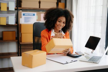 Başlangıçta küçük işletme KOBİ 'si, akıllı telefon ve laptop kullanan girişimci Afrikalı kadın ev ofisinde paket ürün kutularını hazırlamak için online alışveriş siparişi alıyor ve kontrol ediyor
