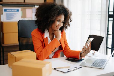 Başlangıçta küçük işletme KOBİ 'si, akıllı telefon ve laptop kullanan girişimci Afrikalı kadın ev ofisinde paket ürün kutularını hazırlamak için online alışveriş siparişi alıyor ve kontrol ediyor