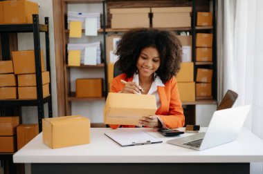 Başlangıçta küçük işletme KOBİ 'si, Girişimci sahibi Afrikalı kadın dizüstü bilgisayar kullanıyor ve paket ürün kutularını hazırlamak için online alışveriş siparişlerini kontrol ediyor. ev ofisinde.