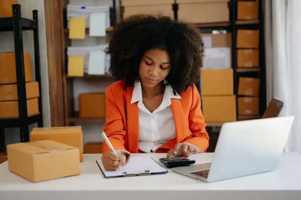 stock image Startup small business SME, Entrepreneur owner African woman using laptop and checking online purchase shopping order to prepare pack product boxes. at home office