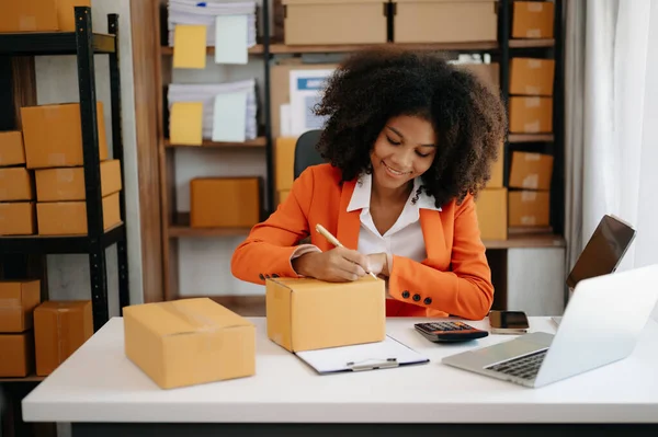 Başlangıçta küçük işletme KOBİ 'si, akıllı telefon ve laptop kullanan girişimci Afrikalı kadın ev ofisinde paket ürün kutularını hazırlamak için online alışveriş siparişi alıyor ve kontrol ediyor