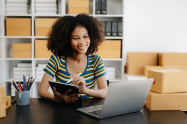 Küçük işletme girişimcisi KOBİ serbest çalışan Afrikalı kadın ev ofisinde kutular, tablet ve dizüstü bilgisayarla, e-ticaret konseptiyle çalışıyor