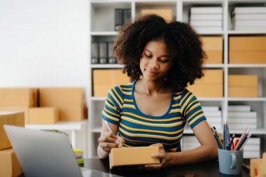 Küçük işletme girişimcisi KOBİ Serbest çalışan Afrikalı kadın ev ofisinde kutular ve laptopla çalışıyor, e-ticaret konsepti