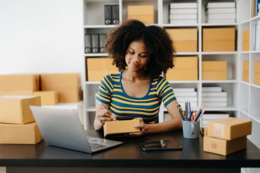 Küçük işletme girişimcisi KOBİ serbest çalışan Afrikalı kadın ev ofisinde kutular, tablet ve dizüstü bilgisayarla, e-ticaret konseptiyle çalışıyor