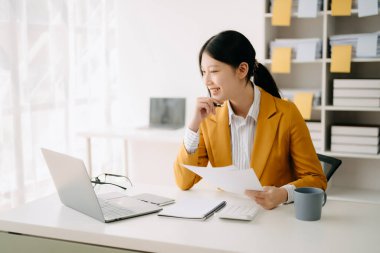 Modern ofiste çalışan, dizüstü bilgisayarı ve telefonu olan genç ve çekici Asyalı kadın. 