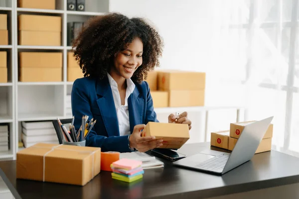 Startup small business SME, Entrepreneur owner African woman using laptop taking receive and checking online purchase shopping order to prepare pack product boxes in office
