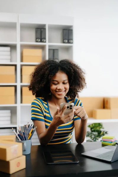 Stock image Small business entrepreneur SME freelance African woman working at home office with boxes and laptop with phone, e-commerce concept