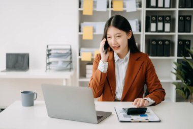 Mutlu Asyalı iş kadını modern ofisteki bilgisayarla akıllı telefondan konuşma zevkine sahip.