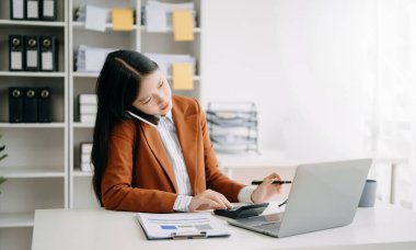 Mutlu Asyalı iş kadını modern ofisteki bilgisayarla akıllı telefondan konuşma zevkine sahip.