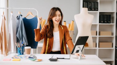 Moda tasarımcısı, genç Asyalı kadın atölyede otururken tablet kullanıyor ve gülümsüyor.