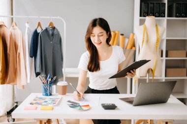 Asyalı moda tasarımcısı masa başında giysiler, hesap makinesi ve dizüstü bilgisayar üzerinde çalışıyor