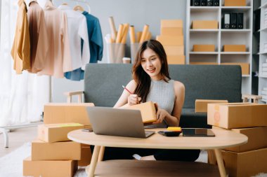 Asyalı bir kadın dizüstü bilgisayarla masa başında çalışıyor ve kargo ve online alışveriş için kutu hazırlıyor. 