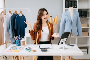 Asyalı moda tasarımcısı kadın modern stüdyoda dijital tablet kullanarak iletişim kuruyor. Giysileri sabah ışığında raflarda asılı duruyor.