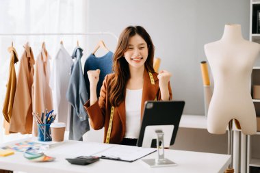 Moda tasarımcısı, genç Asyalı kadın atölyede otururken tablet kullanıyor ve gülümsüyor.