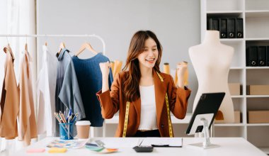 Masada dijital tabletle çalışan Asyalı moda tasarımcısı kadın. Mutluluğu elleriyle işaret ediyor.