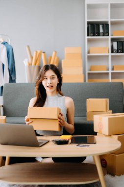 Asyalı bir kadın dizüstü bilgisayarla masa başında çalışıyor ve kargo ve online alışveriş için kutu hazırlıyor. 