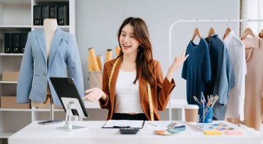 Asyalı moda tasarımcısı kadın modern stüdyoda dijital tablet kullanarak iletişim kuruyor. Giysileri sabah ışığında raflarda asılı duruyor.