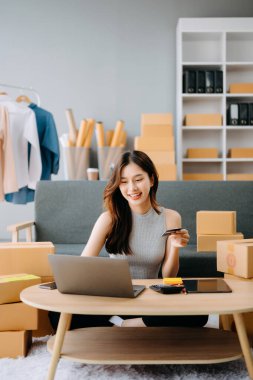 Bilgisayarla masa başında çalışan Asyalı moda tasarımcısı kadın, internette kart ödemeleri yapıyor. 