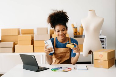 Girişimci sahibi. Akıllı telefon ve tablet kullanan Afrikalı Amerikalı bir kadın paket ürün kutularını hazırlamak için online alışveriş siparişlerini alıyor ve kontrol ediyor. 